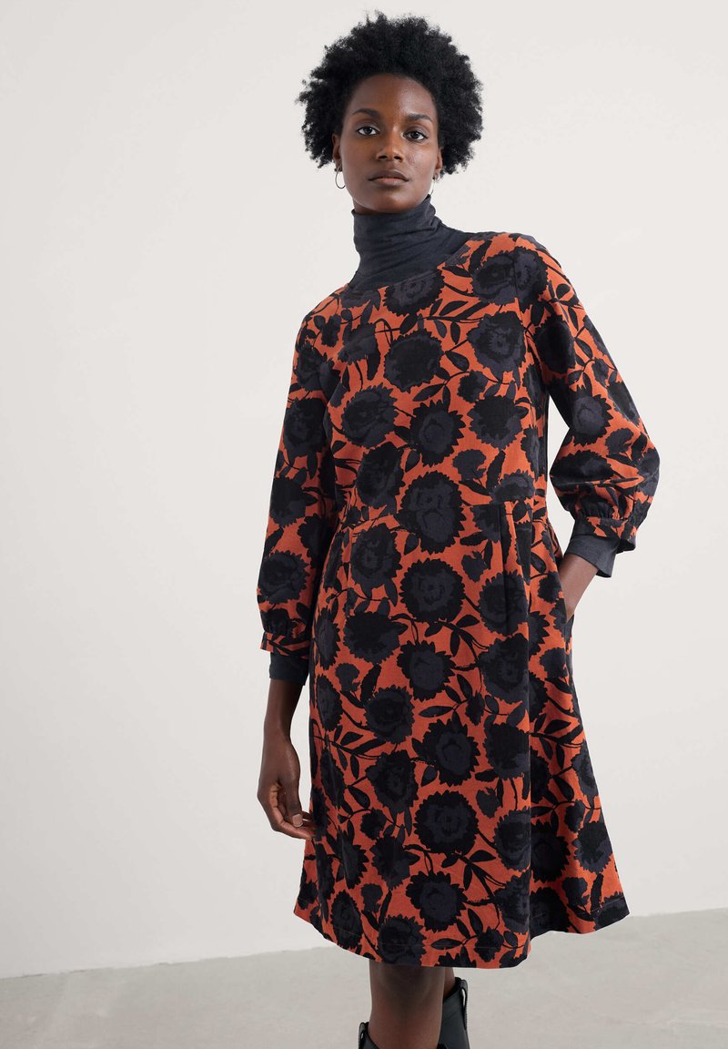 Vestido de manga larga con cuello alto, que presenta un patrón floral negro sobre un fondo naranja. Confeccionado en una tela texturizada, con bolsillos laterales.