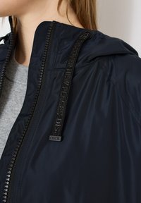 Gros plan sur une personne portant une veste à capuche bleu marine avec une fermeture éclair noire et un cordon noir étiqueté "CECIL".