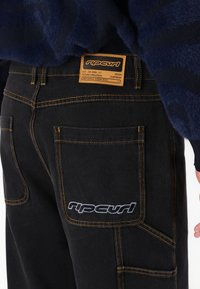 Sorte denimbukser med kontrastfarvet orange syninger, store lommer og et beige mærke med brandnavn og detaljer.