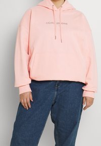 Sudadera rosa claro de algodón. Presenta un bolsillo frontal tipo pouch, cordones y un pequeño logo en el pecho. Combinada con jeans azul oscuro.