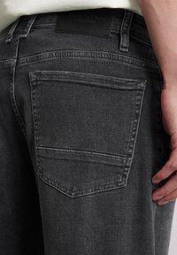 Close-up van een persoon die zwarte denim jeans draagt met een zichtbare achterzak en een hand die vlakbij de dij rust.
