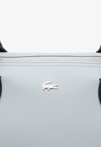 Borsa a mano di colore azzurro chiaro con finiture nere, hardware in argento e un piccolo logo di coccodrillo vicino alla cerniera. Finitura liscia, chiusura a zip.