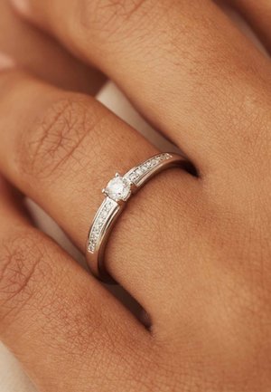 Bague en argent ornée d'un diamant rond central avec de petites pierres transparentes le long de l'anneau, posée sur une surface de peau texturée.