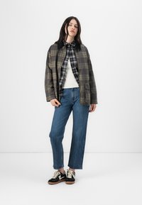 Karirana overshirt u zelenoj i smeđoj boji, složena preko crno-bijele karirane košulje, u kombinaciji s plavim trapericama i crnim tenisicama.