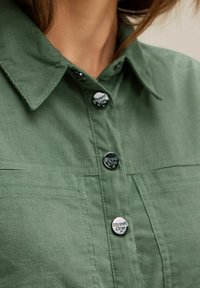 Groene corduroy blouse met een puntige kraag, knoopsluiting aan de voorkant en twee borstzakken. Opvallende details zijn onder andere logoknopen en een gestructureerde stof.