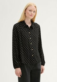 Camisa negra de manga larga con lunares crema, con botones en la parte delantera, cuello y detalle fruncido en los hombros. Se lleva con pantalones oscuros.