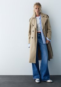 Kvinde iført beige trenchcoat, hvid skjorte, blåstribet skjorte, vide jeans og sølvfarvede sneakers, stående op ad en hvid væg.