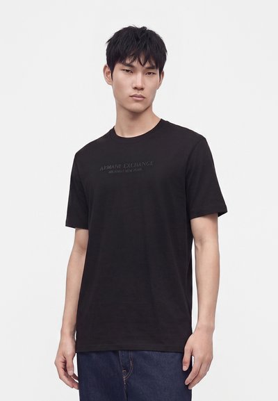 Млад мъж с къса черна коса, облечен в черна тениска Armani Exchange и тъмносини дънки на едноцветен бял фон.