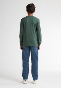 Camicia verde in cotone a maniche lunghe, taglio dritto, colletto tondo, indossata con jeans in denim blu e sneakers bianche. Design semplice, senza motivi visibili.