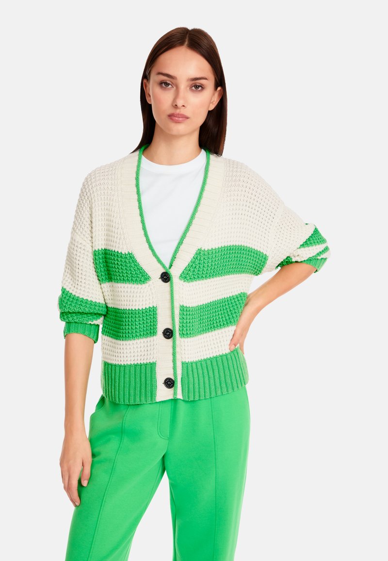 Häkel-Cardigan in Creme- und Grüntönen mit Streifen. Verfügt über einen V-Ausschnitt, eine Knopfleiste und einen gerippten Saum. Wird mit passenden grünen Hosen getragen.