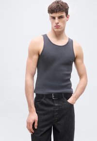 Νέος άνδρας που φοράει σκούρο γκρι αμάνικο τοπ και μαύρα τζιν με ζώνη, στέκεται μπροστά σε απλό ανοιχτό γκρι φόντο.