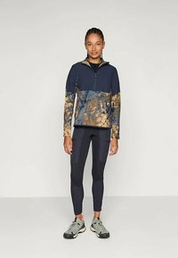 Granatowy sportowy pulower z zamkiem z przodu i złoto-brązowymi fakturowanymi akcentami; w połączeniu z dopasowanymi czarnymi legginsami i szarymi butami do biegania.