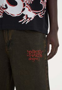 Camiseta gráfica negra con diseño de dragón rojo, combinada con pantalones de mezclilla oscuros que presentan el logo bordado en rojo de "Diseños de Don Ed Hardy" en el bolsillo.