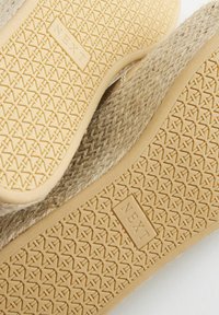Semelles de chaussures beiges texturées avec motif répétitif « X » et logo « NEXT », associées à une tige en fibres naturelles tissées.