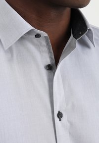 Vaaleanharmaa pukupaita mustakuvioisella kauluksella, jossa on mustat napit ja hienovarainen kiilto kankaassa, korostaen siistiä muotoilua.