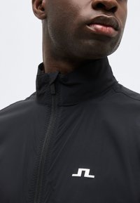 Κοντινό πλάνο ενός ατόμου που φοράει μαύρο μπουφάν με φερμουάρ και ψηλό γιακά, με ένα μικρό λευκό γεωμετρικό λογότυπο στο στήθος.