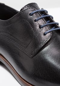 Black leather shoe with a smooth texture, rounded toe, and contrasting blue laces. Features subtle stitching and a sleek design.