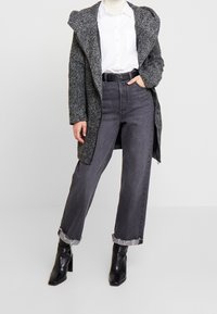 Long manteau gris en tissu texturé, superposé à une chemise blanche, associé à un jean gris taille haute et des bottines noires à talons hauts.