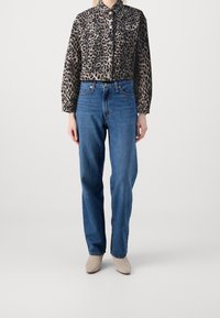En person står självsäkert iförd en jacka med leopardtryck, blå jeans och beige ankellånga stövlar mot en neutral bakgrund.