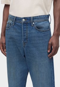 Κοντινό πλάνο ατόμου που φορά μπλε τζιν και μπεζ πουκάμισο, με τα χέρια χαλαρά στα πλευρά και το τζιν κουμπωμένο στη μέση.