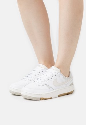 Baskets basses blanches avec le swoosh Nike beige clair portées aux pieds sur un fond blanc uni.