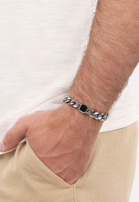 Bracciale in argento con centro rettangolare in pietra nera, indossato su un polso, con tessuto texturizzato e una tasca beige chiaro.