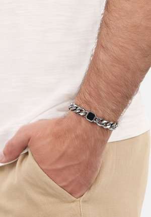 Bracciale in argento con centro rettangolare in pietra nera, indossato su un polso, con tessuto texturizzato e una tasca beige chiaro.