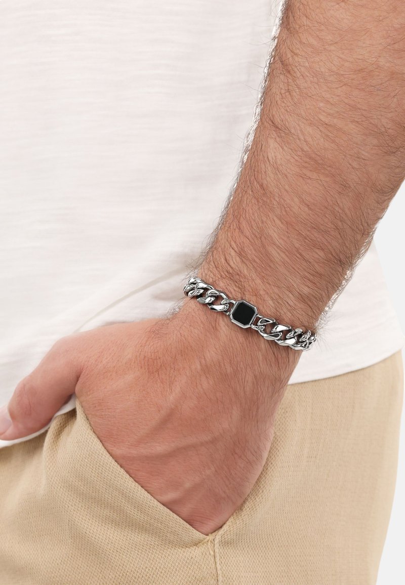 Bracciale in argento con centro rettangolare in pietra nera, indossato su un polso, con tessuto texturizzato e una tasca beige chiaro.