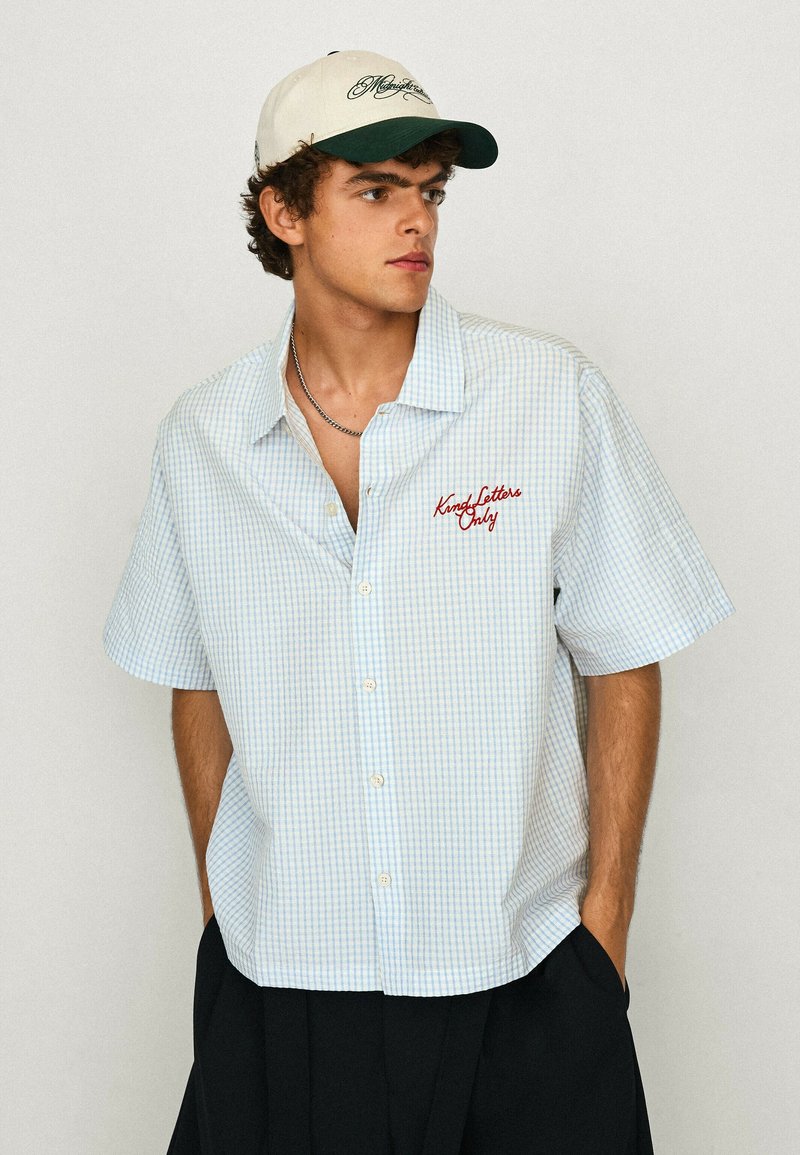 Jeune homme portant une chemise à manches courtes à carreaux blancs et bleus avec un texte rouge et une casquette de baseball blanche, regardant sur le côté, les mains dans les poches.