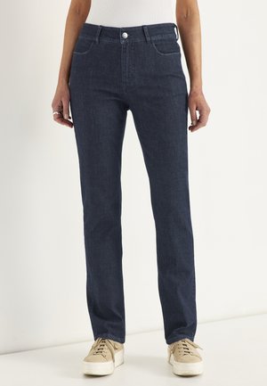 Person iført mørkeblå jeans med lige ben og beige platformsneakers står mod en ensfarvet lys baggrund.