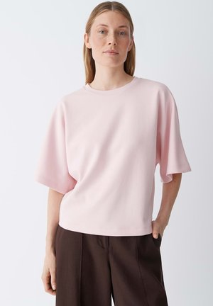 Femme aux cheveux raides portant un haut ample à manches courtes rose clair et un pantalon marron foncé, debout avec une main dans la poche sur fond blanc.