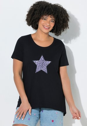 Donna con capelli ricci sorride indossando una t-shirt nera con una stella viola a stampa leopardata e pantaloncini di jeans con piccole toppe a forma di stella.