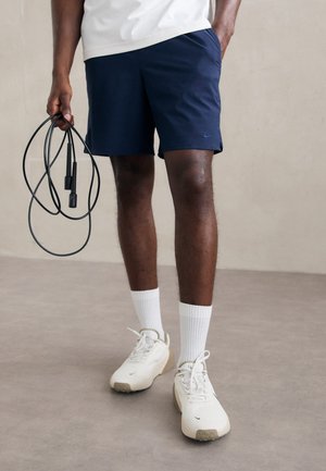 Shorts de sport bleu marine avec poches, chaussettes blanches mi-mollet et baskets blanc cassé. Une corde à sauter noire est tenue dans la main gauche.
