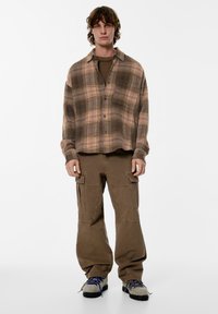 Rjava srajca na karirasto z gumbe z svetlo roza poudarki, kombinirana z rjavimi cargo hlačami z žepi ter sivimi čevlji z modrimi vezalkami.