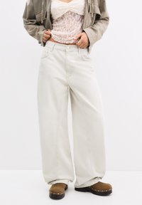Krämfärgade vida jeans med knappframsida, femfickorsdesign och synliga sömnadsdetaljer, ihop med en spetsblus och bruna träskor.