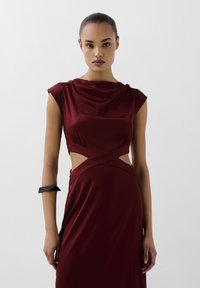 Bourgogne satin kjole med draperet halsudskæring, udskårne sider og en tætsiddende silhuet. Sort geometrisk armbeslag accessory.