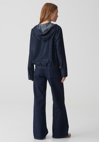 Giacca di jeans con cappuccio, caratterizzata da una vestibilità comoda, orlo elastico e polsini con bottoni. Abbinata a pantaloni in denim a gamba larga, entrambi in blu scuro.