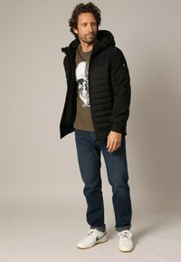 Veste rembourrée noire avec capuche, portée sur un t-shirt graphique marron avec un crâne. Associée à un jean bleu et des baskets blanches.