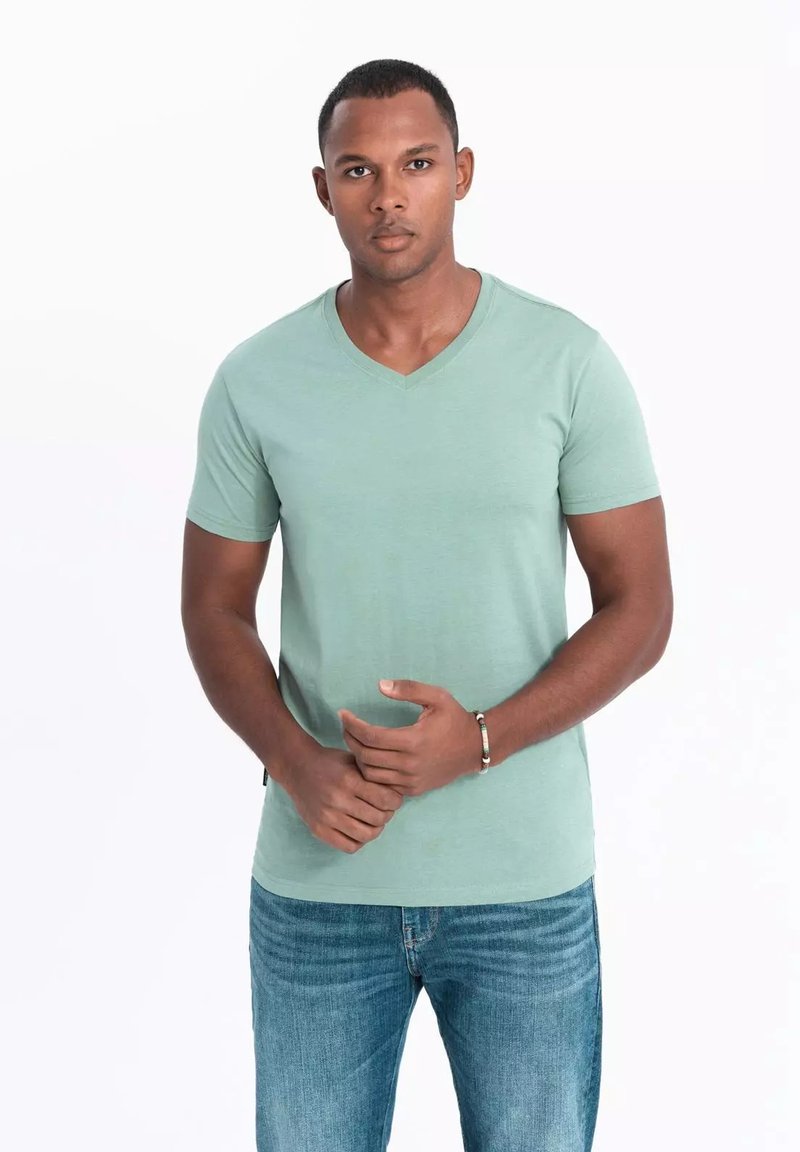 Camiseta verde menta con escote en V, hecha de una tela suave, diseño ajustado, mangas cortas, combinada con jeans azul claro. Hombre posa con las manos entrelazadas.