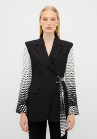 Blazer noir à la silhouette oversize, avec des manches à pois blancs, une ceinture sur le côté et une texture lisse.