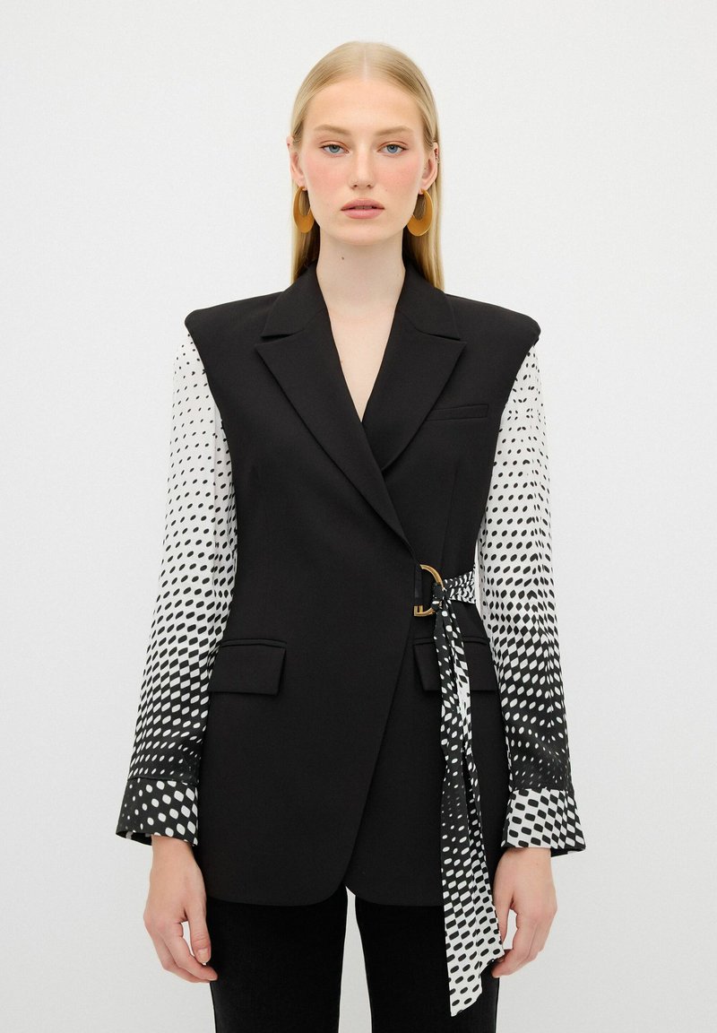 Blazer noir à la silhouette oversize, avec des manches à pois blancs, une ceinture sur le côté et une texture lisse.