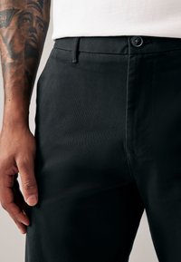 Schwarze Hosen mit maßgeschneiderter Passform, ausgestattet mit einem Knopfverschluss, strukturiertem Stoff und einer glatten Oberfläche, gezeigt zusammen mit einer Hand mit Tätowierung.