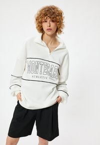 Witte sweatshirt met een half-rits kraag, zwarte "MONTECARLO ATHLETIC" tekst en gestreepte accenten, gecombineerd met zwarte shorts. Zacht materiaal.