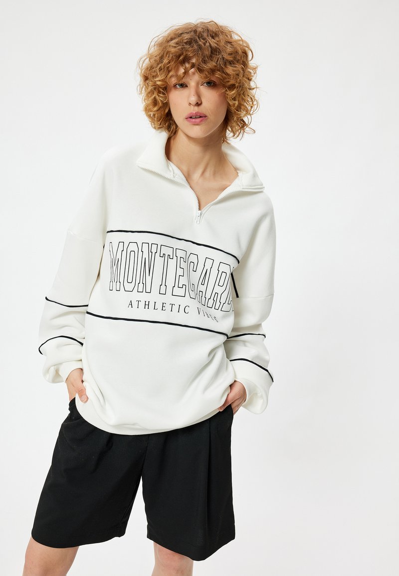 Witte sweatshirt met een half-rits kraag, zwarte "MONTECARLO ATHLETIC" tekst en gestreepte accenten, gecombineerd met zwarte shorts. Zacht materiaal.