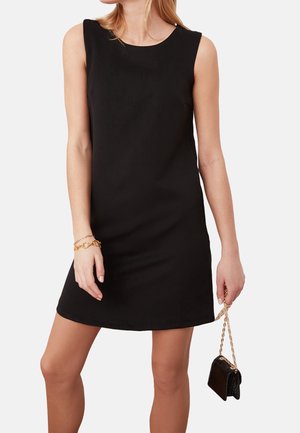 Kobieta ubrana w czarną sukienkę bez rękawów, trzymająca małą czarną torebkę z złotym łańcuszkiem, nosząca złote bransoletki na lewej ręce.