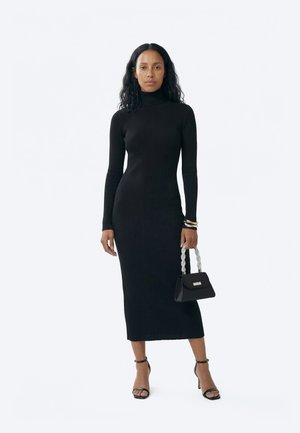Žena s kovrčavom crnom kosom, odjevena u dugu crnu rebrastu haljinu s visokim ovratnikom, crne štikle, zlatne narukvice, držeći crnu torbicu s naramenicom od perlica.