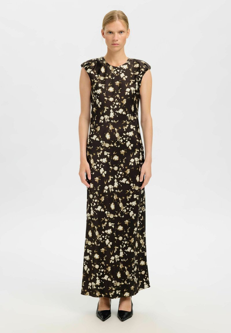 Femme portant une robe maxi noire sans manches avec des motifs floraux beiges, se tenant droite contre un fond clair et uni.