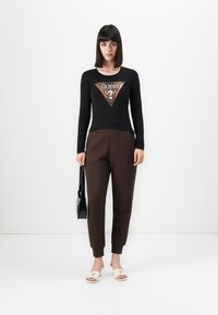 Donna con un taglio di capelli a caschetto nero che indossa un top a maniche lunghe nero con il logo Guess, pantaloni marroni, sandali bianchi e tiene in mano una borsa nera.