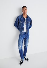 Denim jacket and distressed jeans in deep blue. White T-shirt underneath, black sneakers with white laces, and subtle ripped details on pants.
