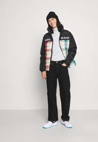 Chaqueta acolchada negra con secciones de cuadros multicolores, logo en el pecho, suéter de cuello alto gris, pantalones cargo negros y zapatillas blancas con detalles azules.